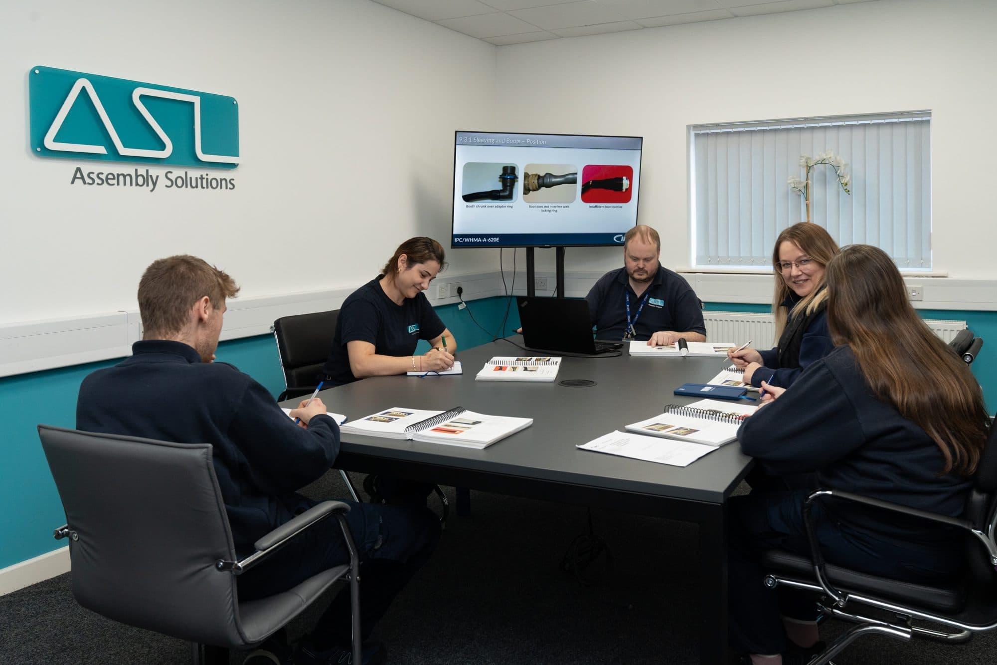 Five members of the ASL team at an IPC/WHMA-A-620 training session around a meeting table, with course workbooks open and a screen showing a slide on sleeving and boot positions, under the Assembly Solutions sign
