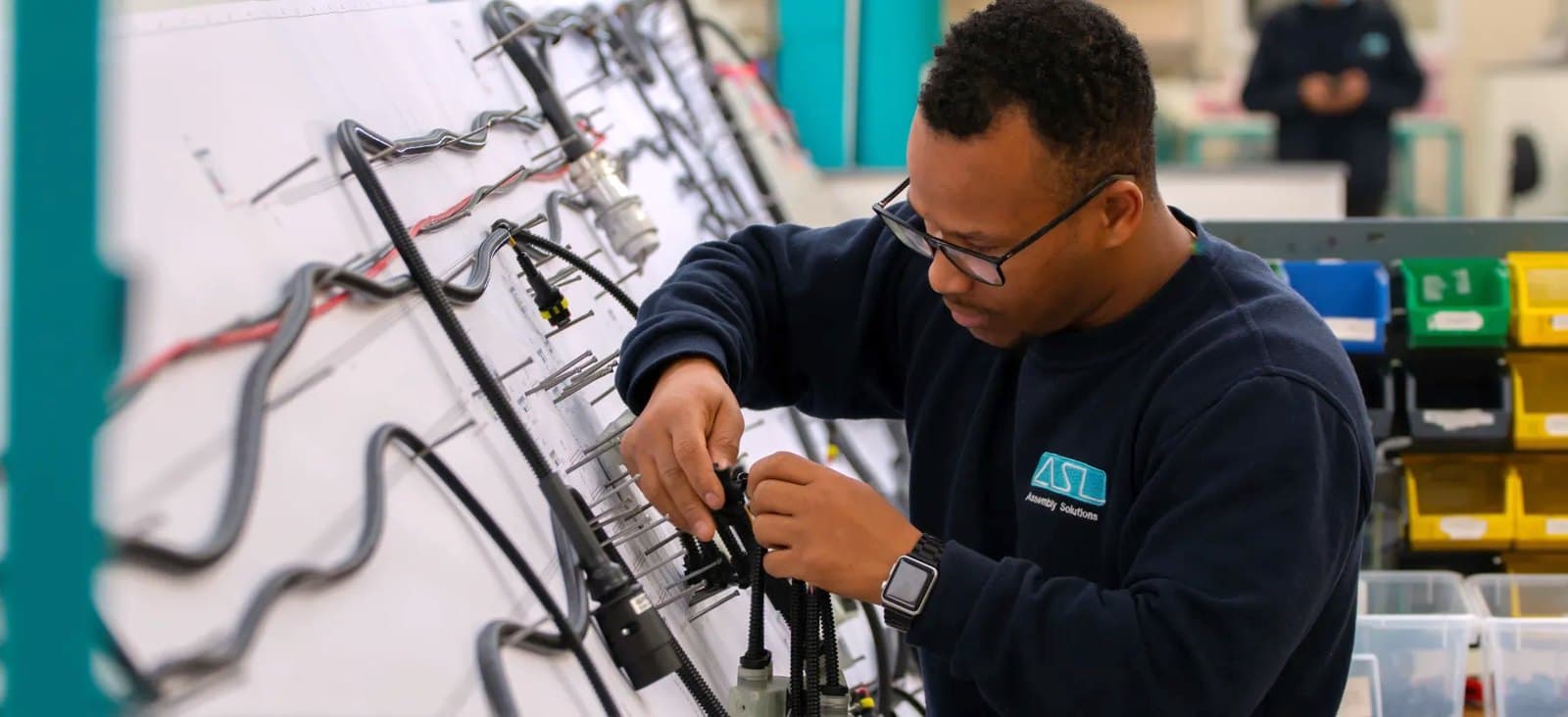 An ASL operator in glasses building a wiring loom on a pegged harness board, with parts bins on the shelves behind