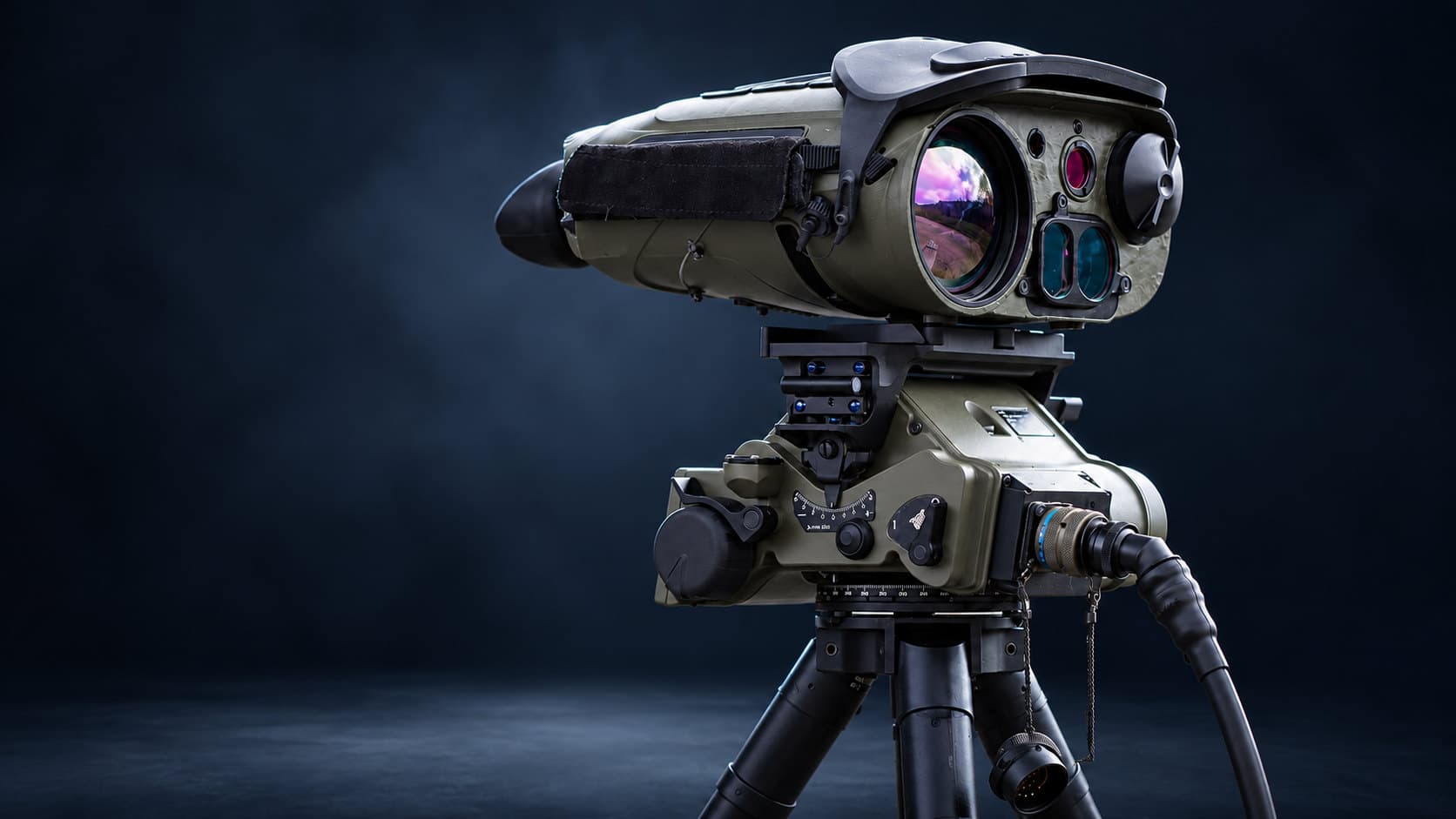 Olive-drab target acquisition unit with a large lens and smaller sensor windows on a tripod mount with angle scales, its black cable joined by an olive-drab circular connector with a blue identification band and a capped connector hanging on a lanyard, against a dark navy background