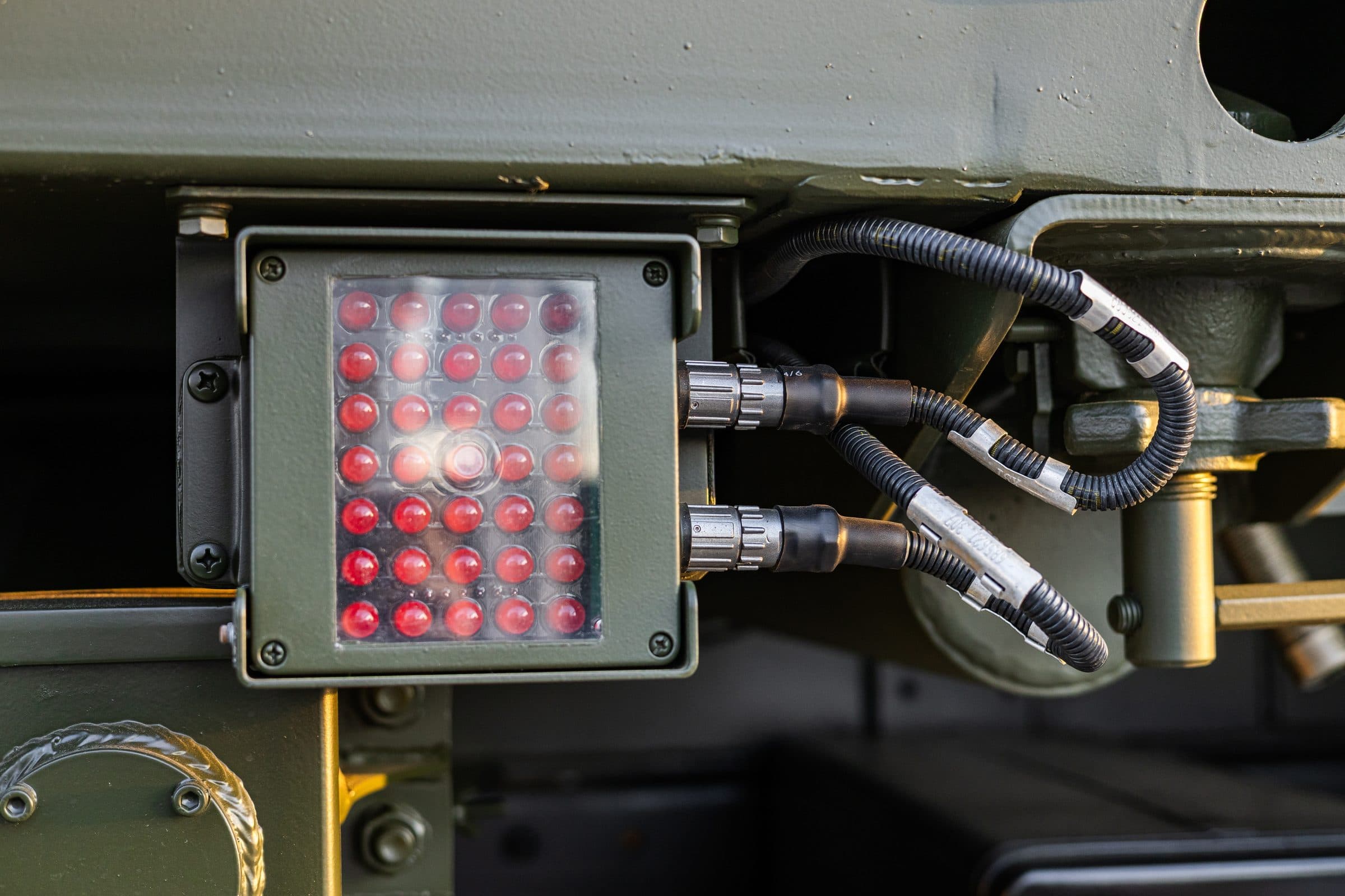 Close-up on an olive-green military vehicle: a red LED light unit with two circular connectors, their black cables in convoluted tubing with marker sleeves